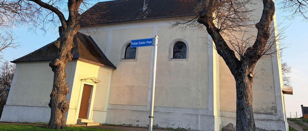 A small church with a steeple and a blue street sign reading 'Pralat Zistler Platz'. The church is surrounded by two bare trees and a clear blue sky.
