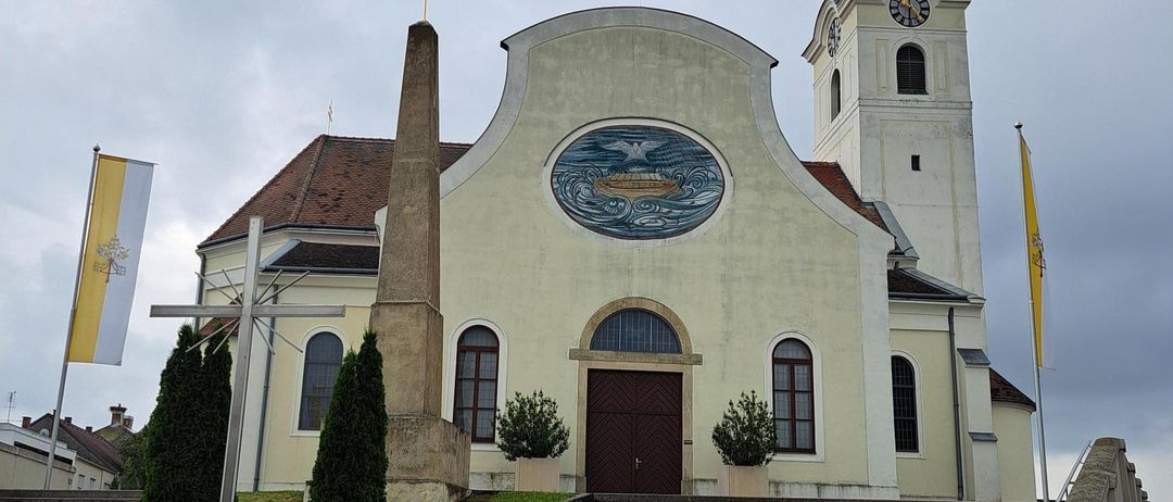 Eine Kirche mit einem großen runden Fenster, einem Kirchturm und einer Uhr. Ein Kreuz und Flaggen sind davor.