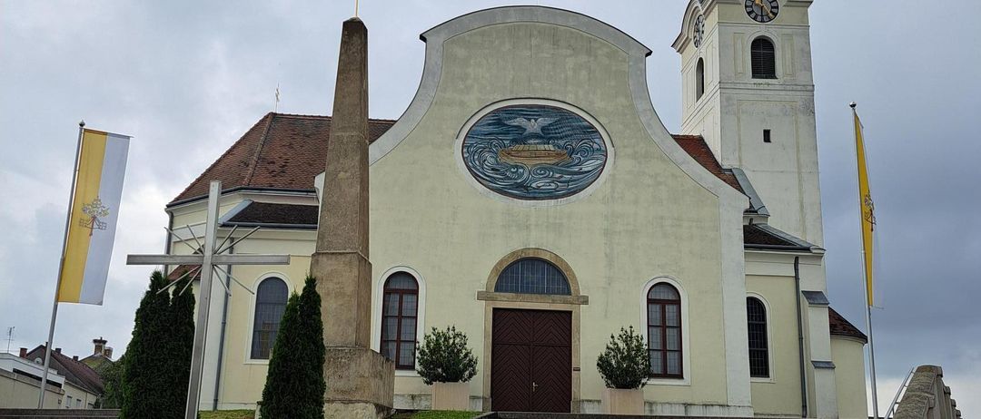 Eine Kirche mit einem Glockenturm und einem Kreuz auf der Spitze, mit einem großen runden Gemälde und einer Treppe, die hinaufführt.