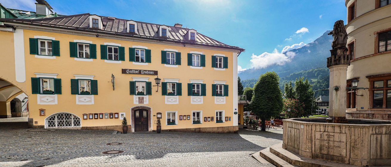 Ein gelbes Gebäude mit grünen Fensterläden und einem Brunnen davor an einem sonnigen Tag mit Bergen im Hintergrund.