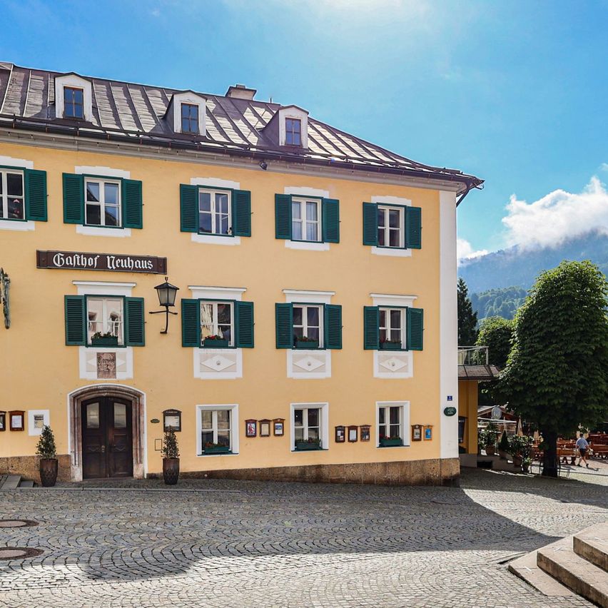 Ein gelbes Gebäude mit grünen Fensterläden und einem Brunnen davor an einem sonnigen Tag mit Bergen im Hintergrund.