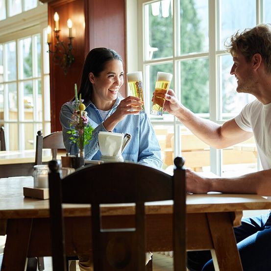 Ein Mann und eine Frau sitzen an einem Tisch in einem Restaurant, lächeln und stoßen mit ihren Biergläsern an.
