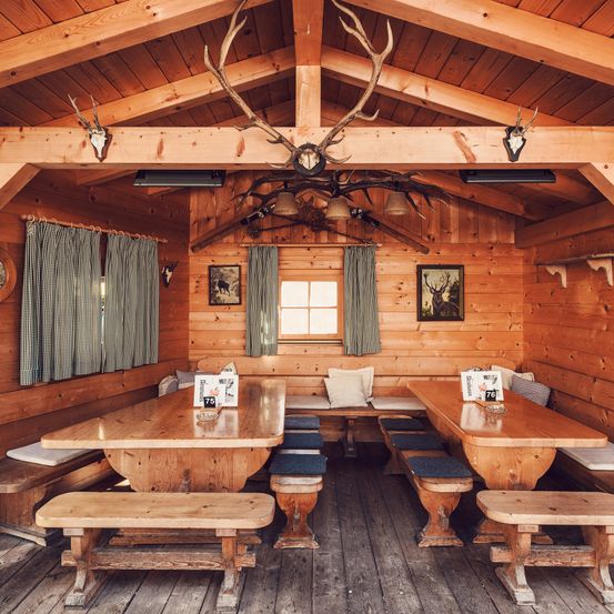 Ein Holzhütten-Interieur mit Bänken, Tischen und Hirschgeweihen an der Wand. Die Hütte verfügt über Holzwände, Vorhänge und ein Fenster.