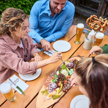 Eine Gruppe von Menschen sitzt an einem Holztisch und genießt eine Mahlzeit mit verschiedenen Gerichten. Die Frau auf der linken Seite hält eine Gabel, und der Mann in der Mitte lächelt. Es gibt Teller, Gläser und Brezeln auf dem Tisch.
