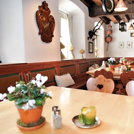 Ein gemütliches Restaurant mit Holzmöbeln, weißen Wänden und einer Blumenarrangement. Herzförmige Stühle, Kerzen und Lampen schmücken den Raum.