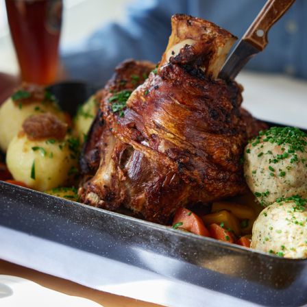 Ein gebratenes Schweineknochen mit Messer, serviert mit Kartoffeln, Karotten und Knödeln. Ein Glas Bier im Hintergrund.