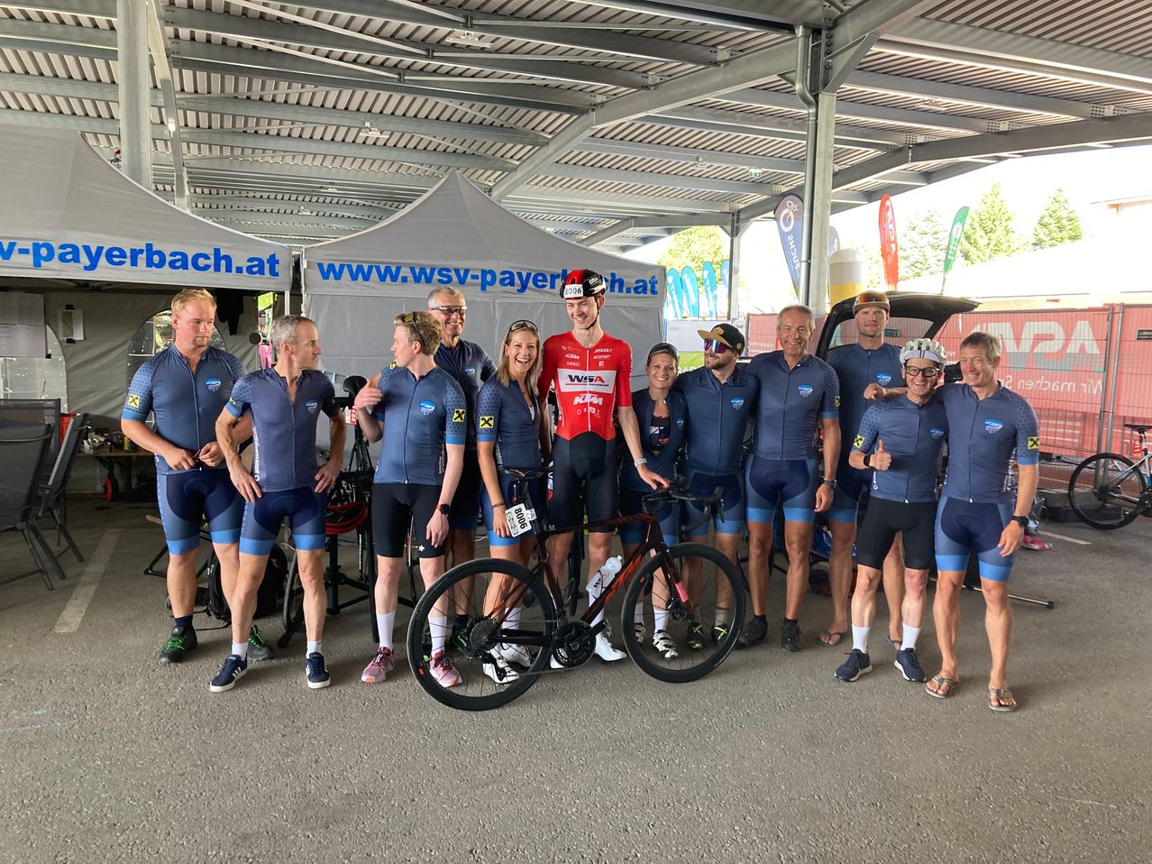 Eine Gruppe von Radfahrern in identischen Uniformen posiert unter einem Zelt mit der Website 'www.wsv-payer.at'. Ein Radfahrer in einem roten Trikot steht neben einem Fahrrad.