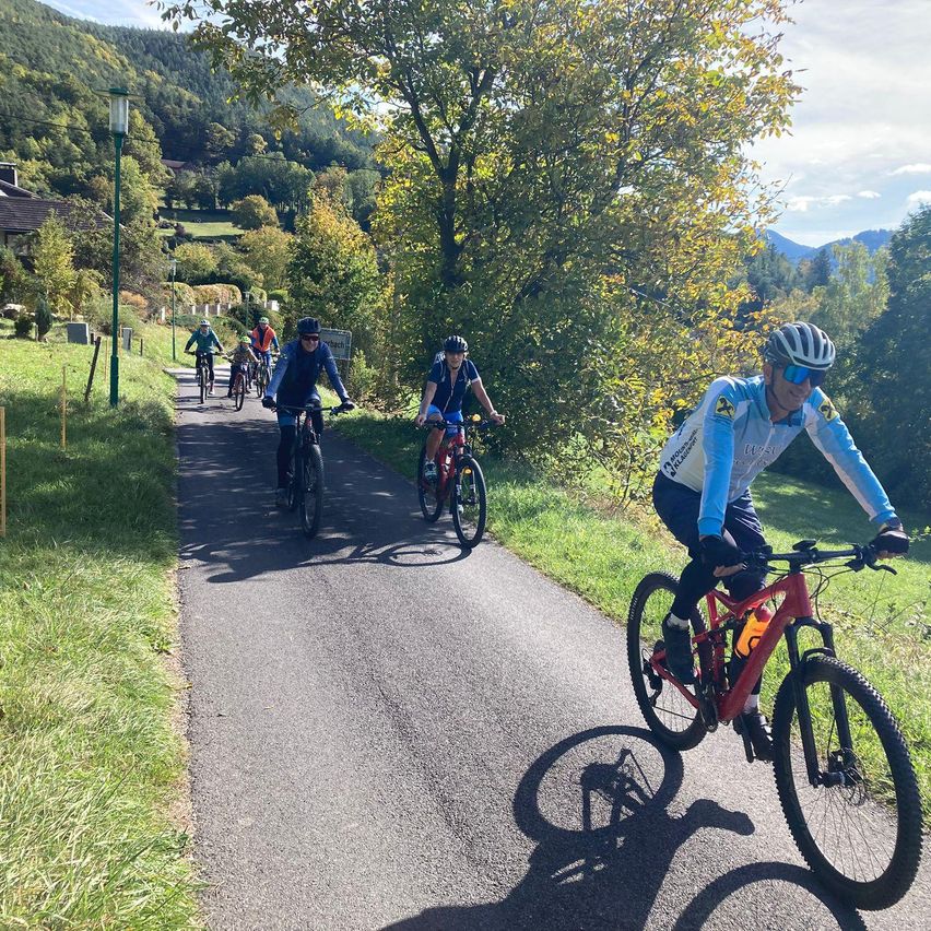 Eine Gruppe von Radfahrern fährt auf einem schattigen Pfad mit Bergen im Hintergrund und trägt Helme und Fahrradausrüstung.