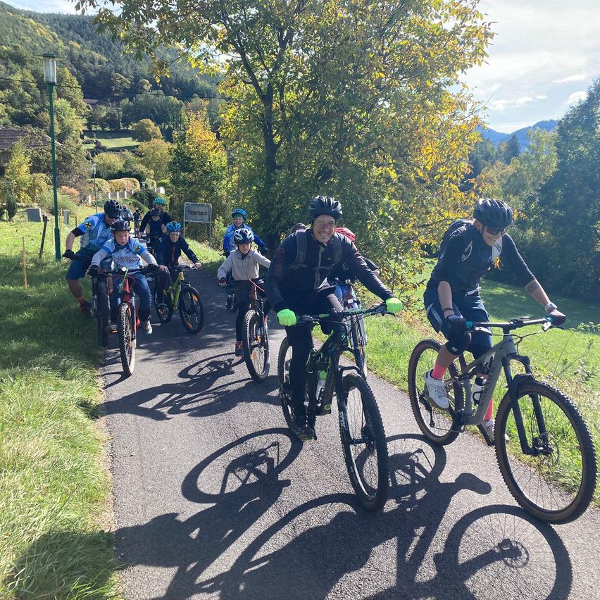 Eine Gruppe von Radfahrern mit Helmen fährt auf einer Straße mit Gras auf beiden Seiten, einem Baum und Bergen in der Ferne.