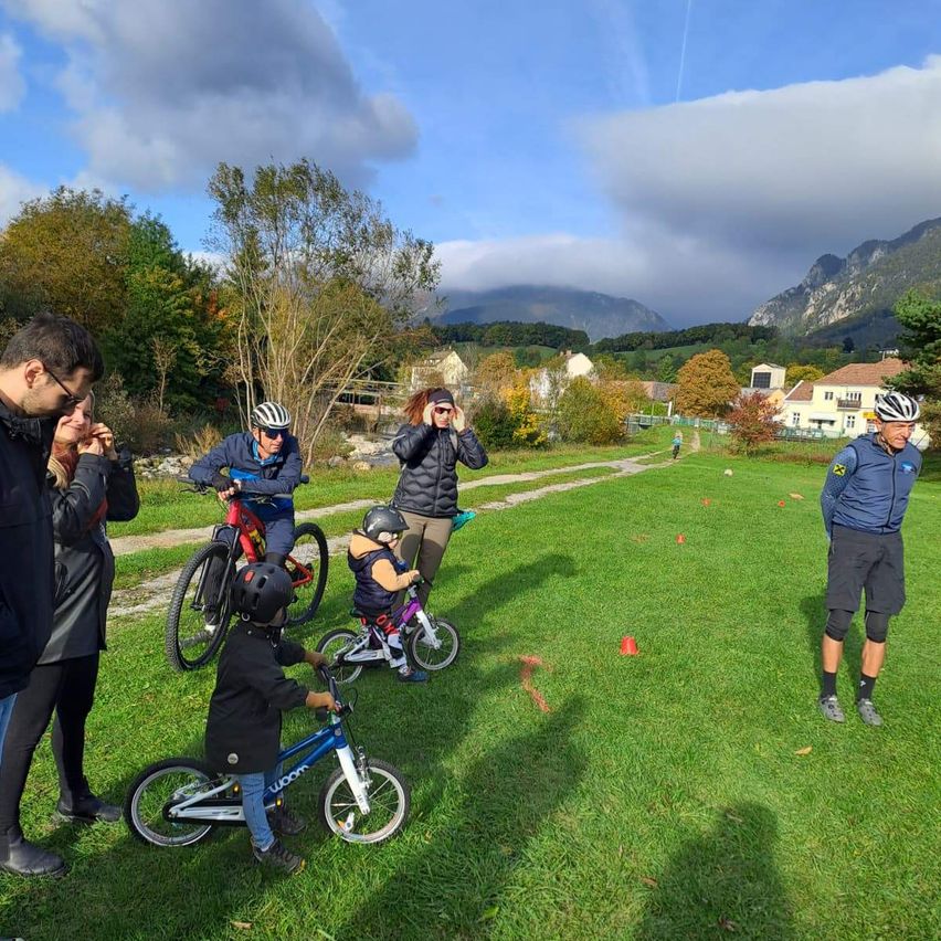 Eine Gruppe von Menschen, darunter Kinder, fahren auf einem Rasenfeld mit Bergen im Hintergrund Fahrräder.
