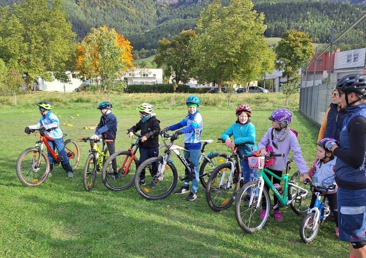 Eine Gruppe von Kindern mit Helmen und Handschuhen steht mit ihren Fahrrädern auf einer Wiese. Dahinter sind Bäume und Büsche. In der Ferne befindet sich ein Gebäude und einige Autos, die davor geparkt sind. Der Himmel ist blau.