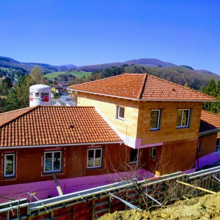 Eine Luftaufnahme eines neuen Hauses im Bau mit einem braunen Dach, Holzwänden und einem weißen Tank oben drauf. Das Gebiet ist umgeben von Bäumen, Bergen und einem klaren blauen Himmel.