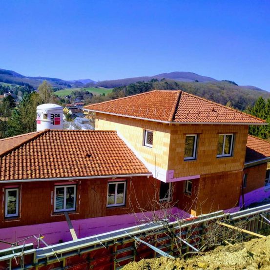 Eine Luftaufnahme eines neuen Hauses im Bau mit einem braunen Dach, Holzwänden und einem weißen Tank oben drauf. Das Gebiet ist umgeben von Bäumen, Bergen und einem klaren blauen Himmel.