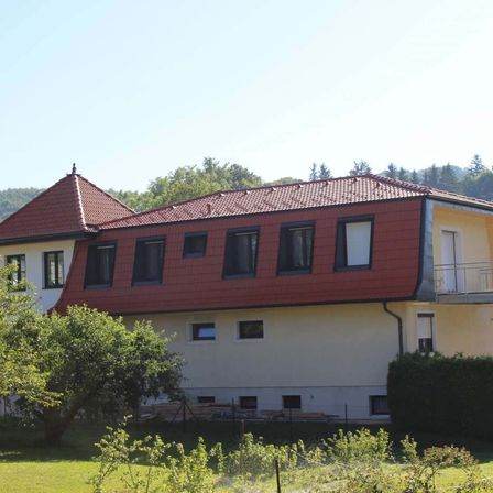 Ein Haus mit rotem Dach und weißen Wänden, umgeben von Grün, steht in einer ländlichen Umgebung mit Bergen im Hintergrund.