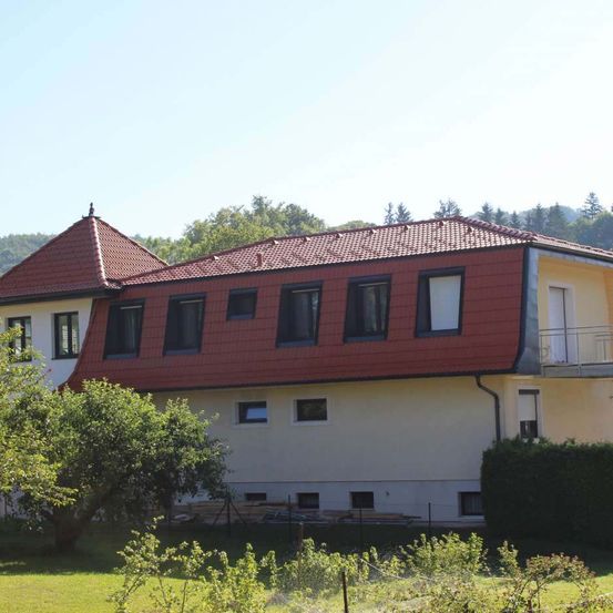 Ein Haus mit rotem Dach und weißen Wänden, umgeben von Grün, steht in einer ländlichen Umgebung mit Bergen im Hintergrund.
