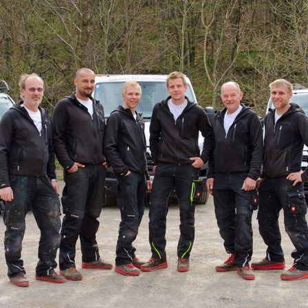 Sechs Männer, die schwarze Jacken und Hosen tragen, stehen nebeneinander auf einem Feldweg mit Fahrzeugen im Hintergrund.