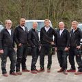 Sechs Männer, die schwarze Jacken und Hosen tragen, stehen nebeneinander auf einem Feldweg mit Fahrzeugen im Hintergrund.
