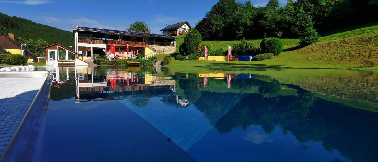 Ein ruhiger Außenpool mit einem Gebäude und Solarpanelen im Hintergrund, umgeben von üppigem Grün und Bergen.