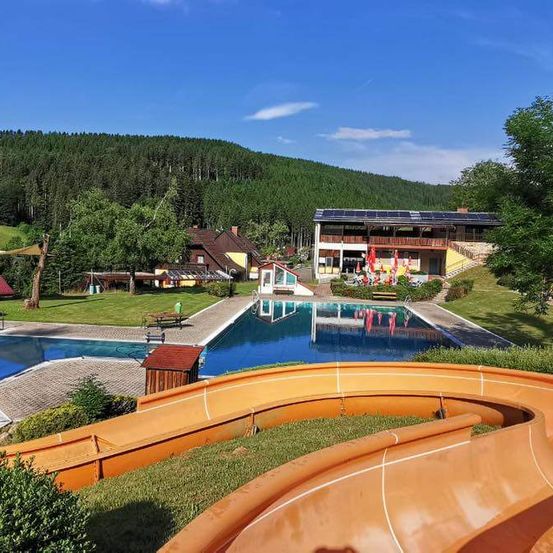 Luftaufnahme eines Resorts mit einem großen Pool, einer Wasserrutsche und Gebäuden mit Solarpanelen. Bäume und Berge im Hintergrund.