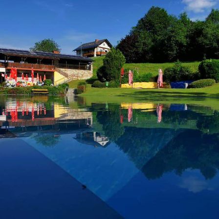 Ein sonniger Tag mit klarem blauem Himmel, einem Gebäude mit Solarmodulen und einem Pool mit Reflexionen von Sonnenschirmen und Bäumen.