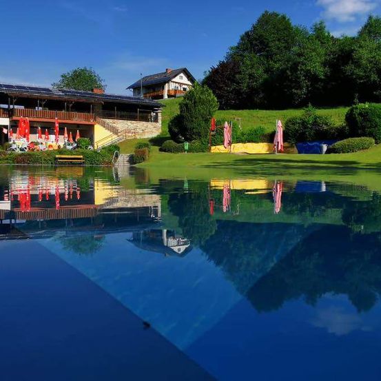 Ein sonniger Tag mit klarem blauem Himmel, einem Gebäude mit Solarmodulen und einem Pool mit Reflexionen von Sonnenschirmen und Bäumen.