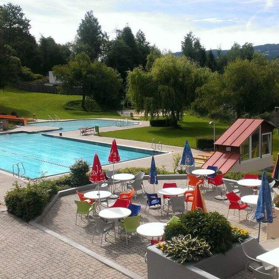 Ein Blick von oben auf einen Außenpoolbereich mit einem großen Schwimmbecken, bunten Tischen und Stühlen und Sonnenschirmen. Der Poolbereich ist von einem Rasenfeld und Bäumen umgeben.