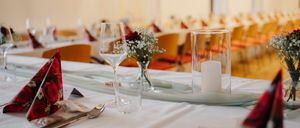 Ein Tisch für ein formelles Abendessen mit weißer Tischdecke, Blumen, Weingläsern und einer Kerze in einem Glashalter. Andere Tische und Stühle sind im Hintergrund.