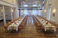Ein langer Bankettsaal mit mehreren für eine Veranstaltung gedeckten Tischen, Holzböden und Glasfenstern mit Blick nach draußen.