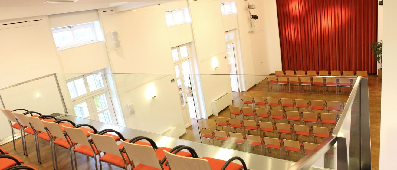 Ein leerer Hörsaal mit roten Vorhängen, Reihen von Stühlen und einem Holzboden. Die Stühle sind in ordentlichen Reihen angeordnet.