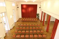 Ein Konferenzraum mit Holzboden und roten Stühlen, die in Reihen angeordnet sind. Es gibt einen roten Vorhang im Hintergrund und eine Pflanze in der Ecke. Der Raum ist leer.