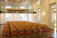 Ein leerer Konferenzsaal mit Reihen von Holzstühlen mit roten Kissen. Links befindet sich ein Balkon mit Stühlen und rechts sind zwei Türen.