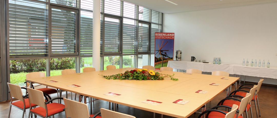 Ein Konferenzraum mit großen Fenstern und einem großen Tisch mit Blumengesteck in der Mitte. Stühle mit roten Kissen umgeben den Tisch. Ein Banner ist an der Wand und ein Projektor befindet sich an der Decke.