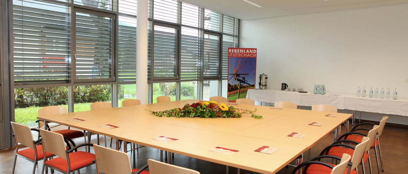 Ein Konferenzraum mit großen Fenstern und einem großen Tisch mit Blumengesteck in der Mitte. Stühle mit roten Kissen umgeben den Tisch. Ein Banner ist an der Wand und ein Projektor befindet sich an der Decke.