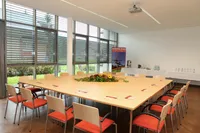 Ein leerer Konferenzraum mit einem langen Holztisch, umgeben von roten Stühlen. Ein Blumenarrangement steht auf dem Tisch. Ein Poster ist an der Wand. Eine Kaffeemaschine steht auf dem Tresen.