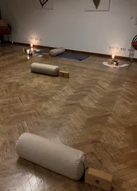 Ein Raum mit Holzboden, mehreren Yoga-Matten und aufgerollten Kissen. Kerzen sind auf dem Boden platziert und ein Stuhl steht in der Nähe der Wand. Es gibt Steckdosen und einen Lichtschalter an der Wand.