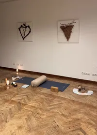 Ein Yoga-Setup auf einem Holzboden mit Yogamatte, gerollter Decke und verschiedenen Gegenständen wie Schüssel, Buch und Kerzen. Wandkunst und Steckdosen sind sichtbar.
