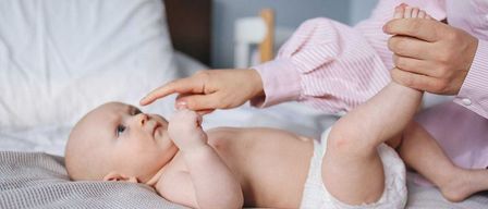 Ein Baby liegt auf einem Bett und trägt eine Windel. Ein Erwachsener in einem rosa gestreiften Hemd hält die Hand des Babys und kitzelt es. Das Baby schaut neugierig auf und der Ort ist drinnen.