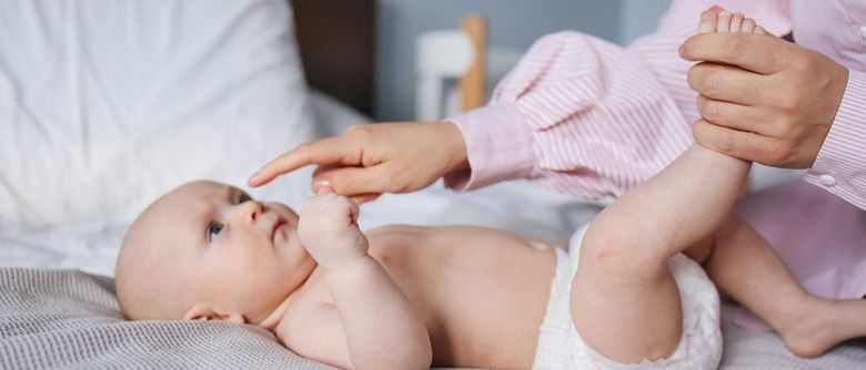 Ein Baby liegt auf einem Bett und trägt eine Windel. Ein Erwachsener in einem rosa gestreiften Hemd hält die Hand des Babys und kitzelt es. Das Baby schaut neugierig auf und der Ort ist drinnen.