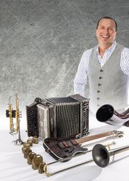 Ein lächelnder Mann in einer Weste steht hinter einem Tisch mit verschiedenen Musikinstrumenten, darunter ein Akkordeon, eine Trompete, ein Tenorhorn und eine Gitarre.