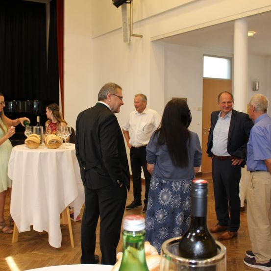 Eine Gruppe von Menschen versammelt sich in einem Raum, wahrscheinlich für ein Geschäftstreffen. Eine Frau steht vor einem Tisch mit einer Flasche Wein und Brot. Die Menschen sind in formeller Kleidung gekleidet.