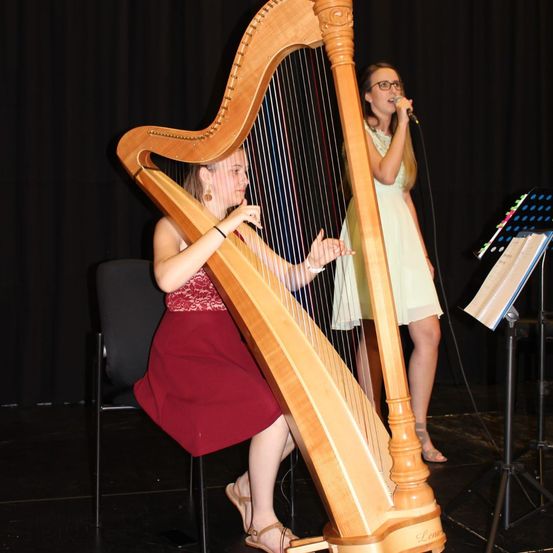 Zwei Frauen auf einer Bühne; eine spielt Harfe und die andere singt mit einem Mikrofon.
