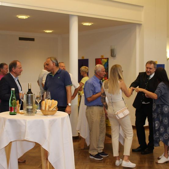 Eine Gruppe von Menschen unterhält sich bei einer Veranstaltung. Ein Tisch mit Weinflaschen, Weingläsern und Brot ist aufgestellt. Der Raum hat weiße Wände, einen Holzboden und eine Decke mit Lichtern.