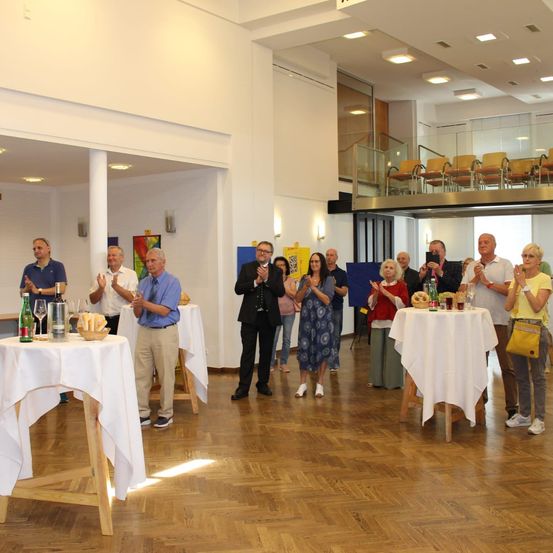 Eine Gruppe von Menschen klatscht in einem Raum mit Tischen und Stühlen. Einige stehen um Tische mit Flaschen, Brot und Weingläsern. Eine Galerie mit Stühlen ist im Hintergrund sichtbar.