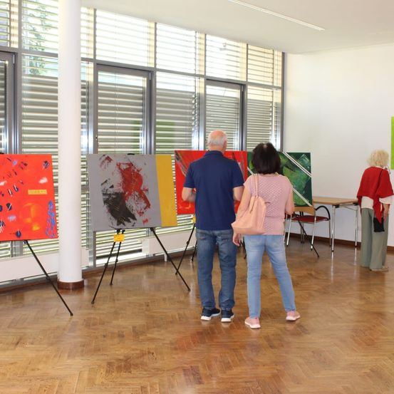Ein Innenbereich einer Kunstgalerie mit mehreren abstrakten Gemälden auf Staffeleien. Ein Paar betrachtet die Gemälde, während eine weitere Person in der Nähe steht. Der Raum hat Holzböden und große Fenster mit Jalousien.