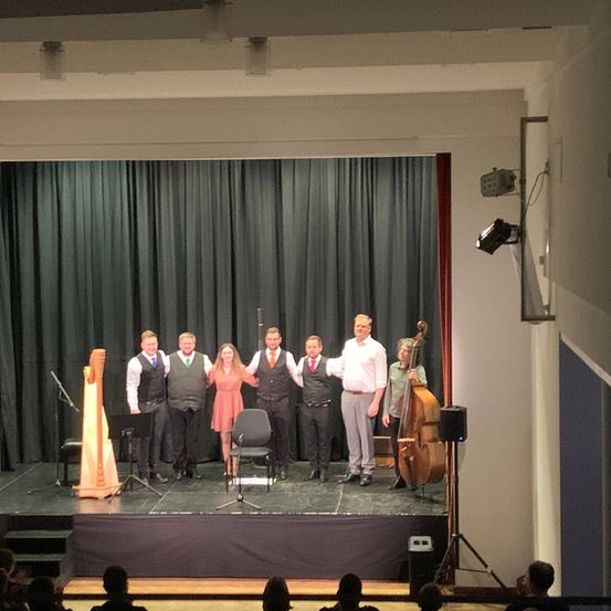 Eine Gruppe von Musikern steht auf der Bühne, darunter eine Harfenistin, ein Cellist und ein Pianist, alle in formeller Kleidung. Sie stehen vor einem schwarzen Vorhang und einigen Musikinstrumenten. Das Publikum sitzt vor der Bühne.