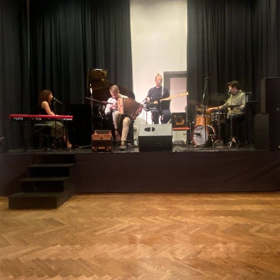 Eine Band spielt auf einer Bühne mit verschiedenen Instrumenten wie Klavier, Gitarre und Schlagzeug. Eine Frau spielt Klavier, während ein Mann Gitarre und ein anderer Akkordeon spielt. Sie alle singen in Mikrofone.