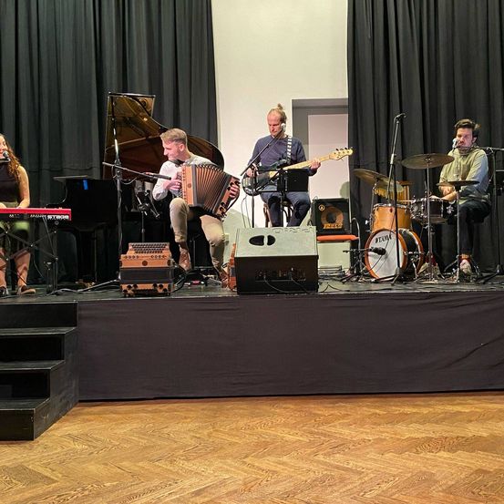 Eine Band spielt auf einer Bühne mit Klavier, Gitarre, Akkordeon und Schlagzeug. Eine Frau spielt links eine Tastatur.