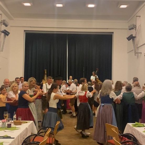 Eine Gruppe von Menschen in traditioneller Kleidung tanzt in einem Raum mit Tischen und Stühlen. Eine Person spielt Cello in der Nähe einer großen schwarzen Leinwand.