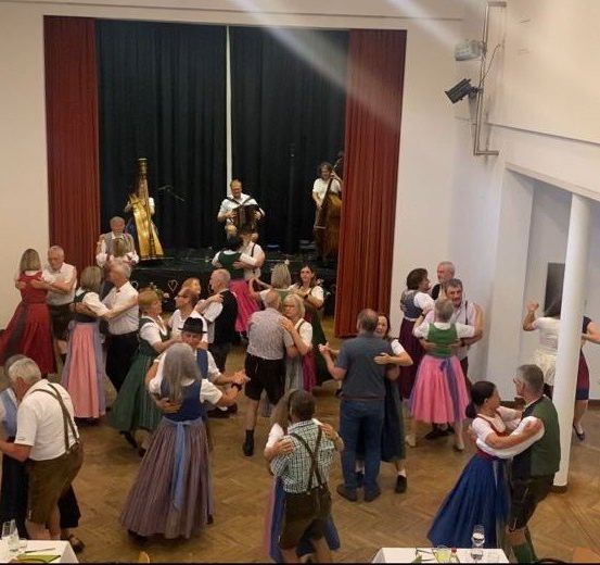 In einem Saal tanzen eine Gruppe von Menschen um eine Bühne, auf der Musiker Instrumente spielen. Die Musiker spielen Akkordeon und Cello.