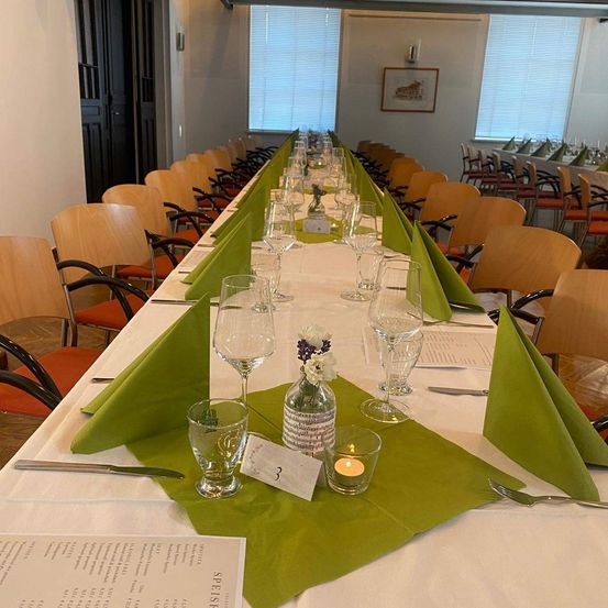 Ein langer Tisch mit weißer Tischdecke und grünen Servietten, Gläsern und Kerzen. Der Tisch ist in einem Esszimmer mit Holzstühlen und einer weißen Wand aufgestellt.